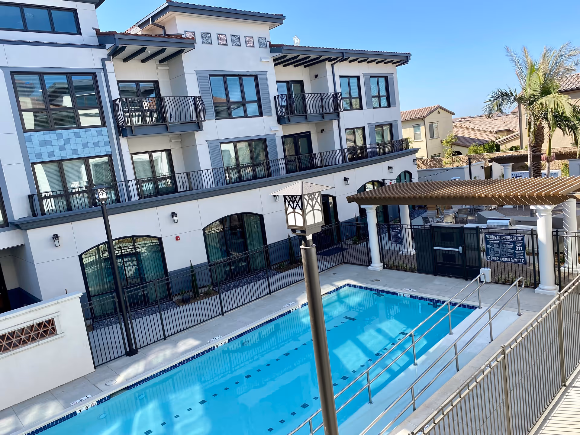 Outdoor swimming pool area at Westmont of Cypress with a fenced pool, a pergola with seating, palm trees, and a multi-story residential building in the background under a clear blue sky.