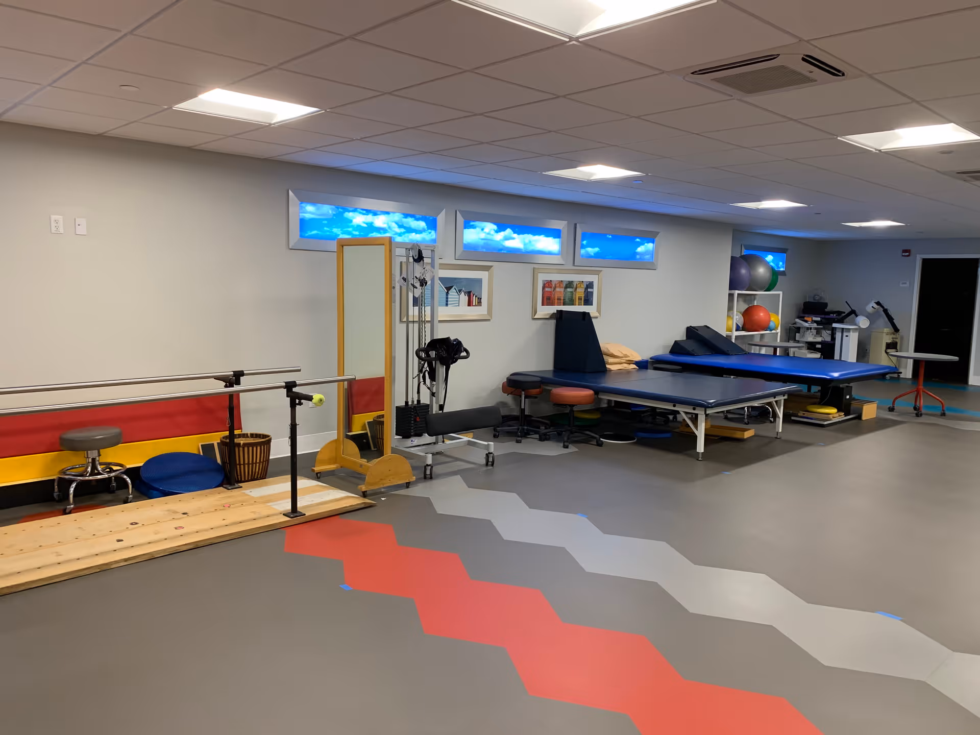 A physical therapy or rehabilitation room with parallel bars for walking exercises, therapy tables, exercise balls, stools, and various therapy equipment. The room has a gray floor with red and gray geometric patterns and small windows near the ceiling showing a blue sky with clouds.