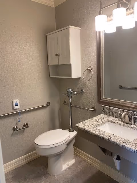 A small bathroom with a white toilet, a wall-mounted cabinet above it, and a granite countertop sink with a large mirror above. The walls are painted beige and there are metal grab bars installed near the toilet for accessibility. A light fixture with three lights is mounted above the mirror.