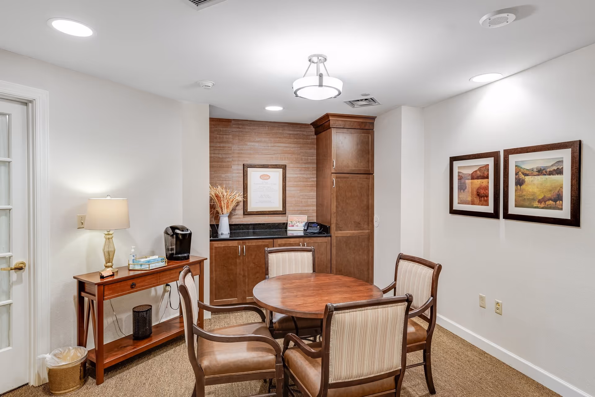 A small, cozy seating area in a senior living facility with a round wooden table surrounded by four cushioned chairs. There is a wooden side table with a lamp, a coffee maker, and a tissue box. The back wall features wooden cabinetry with a framed document and a vase with dried plants. Two framed landscape paintings hang on the adjacent wall. The room is well-lit with ceiling lights.