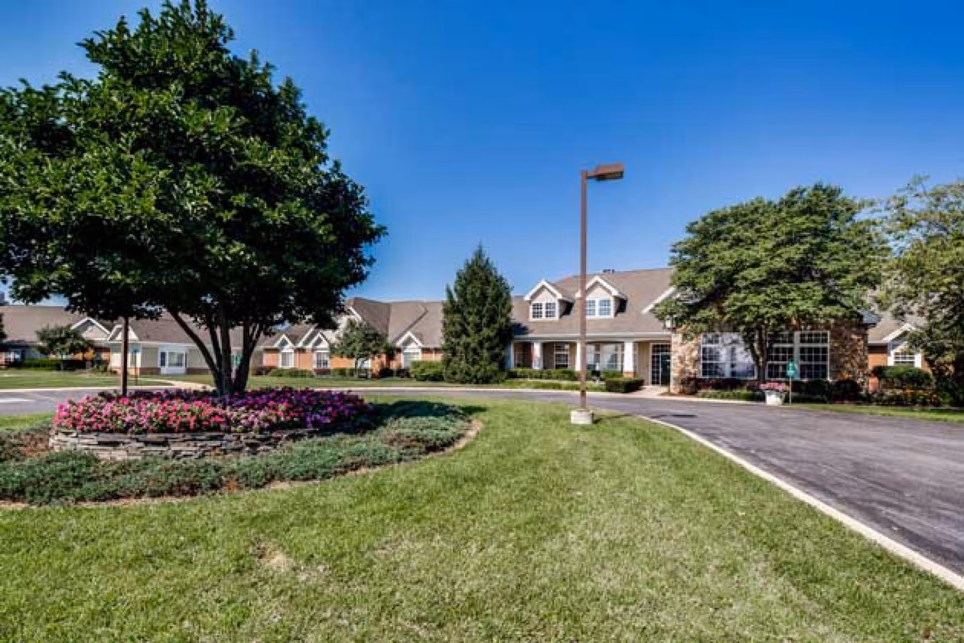 Exterior view of Somerford House And Place Hagerstown showing a well-maintained lawn with a circular flower bed featuring a tree and pink flowers, a paved driveway, and a building with multiple windows and a stone facade under a clear blue sky.