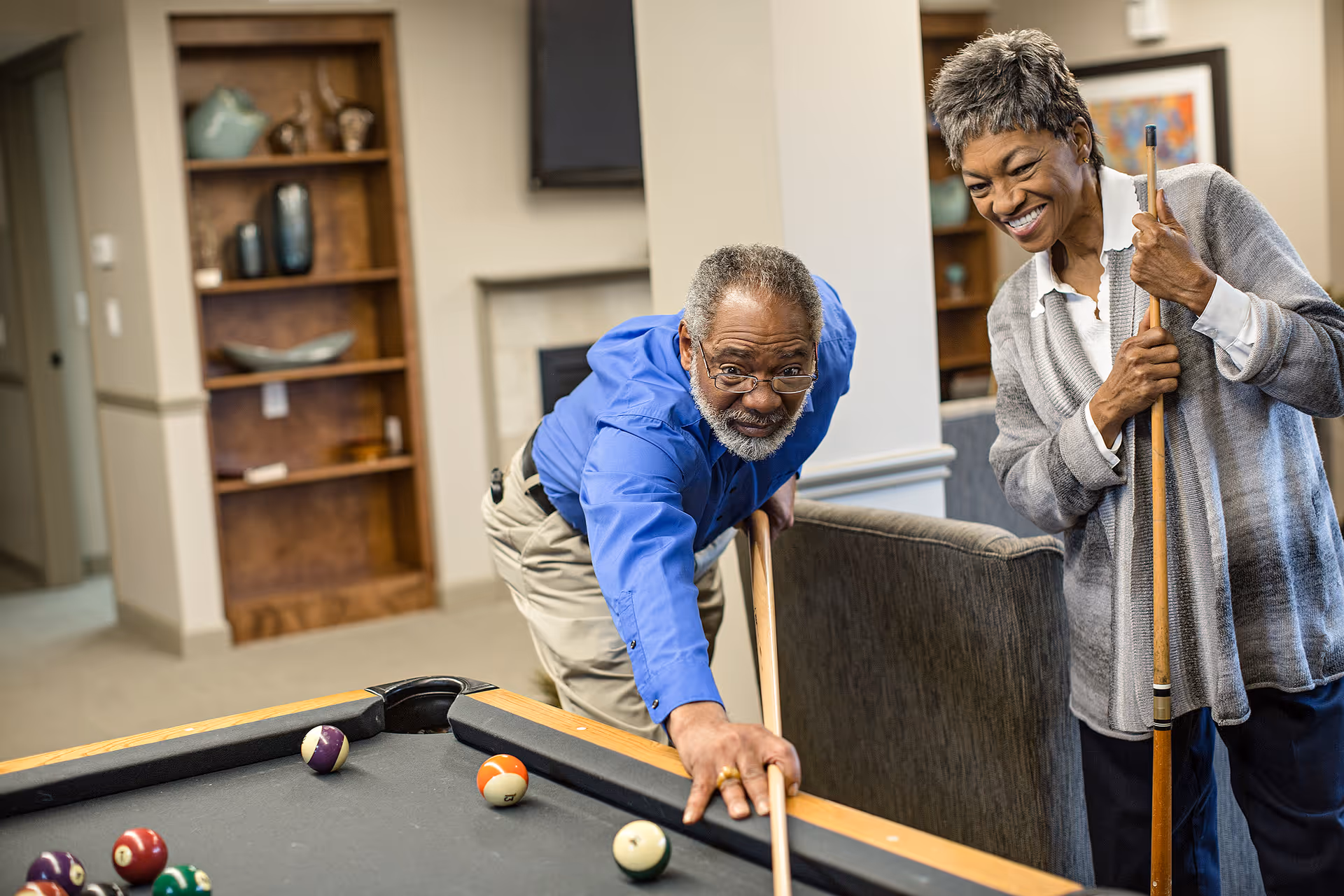 An elderly man and woman playing pool together in a cozy indoor common area. The man is leaning over the pool table aiming a shot with a cue stick, while the woman stands nearby smiling and holding her cue stick. Behind them are shelves with decorative items and a fireplace.
