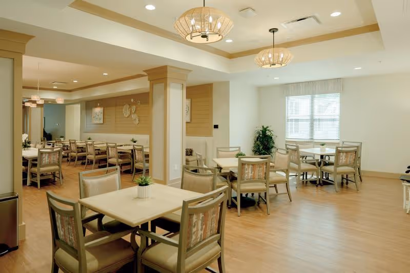 A bright and spacious dining room with several square tables and cushioned chairs arranged neatly. The room features light wood flooring, beige walls with decorative paneling, large windows with blinds allowing natural light, and modern pendant lights hanging from the ceiling. Small potted plants are placed on the tables and in the corners, creating a welcoming atmosphere.