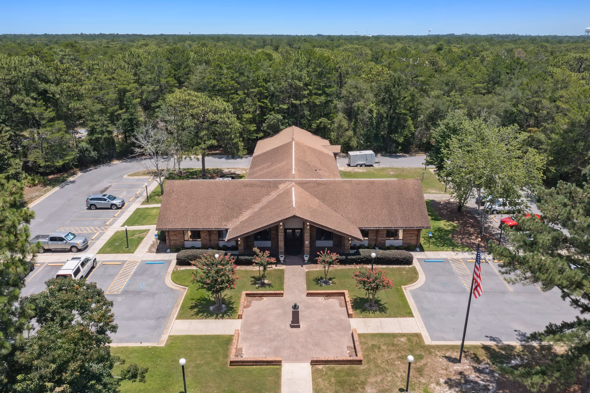 Aerial view of a single-story building with a brown roof surrounded by parking spaces and greenery. The building is centered with a paved walkway leading to the entrance, flanked by small trees and landscaped areas. An American flag is visible on a flagpole to the right side of the building. The background shows a dense forest under a clear blue sky.