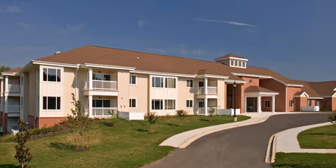 Exterior view of a two-story senior living facility building with beige siding and brick accents, a brown roof, balconies, and a curved driveway leading to the entrance under a clear blue sky.