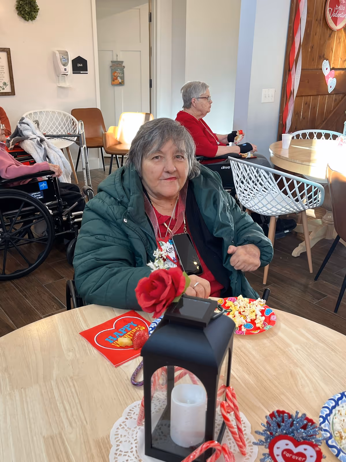 An elderly woman in a green jacket sitting at a wooden table with Valentine's Day decorations, including a red rose and a black lantern with a candle inside. She is in a room with other elderly people, some in wheelchairs, and there are chairs and a round table in the background with more Valentine's Day decorations on the wall.