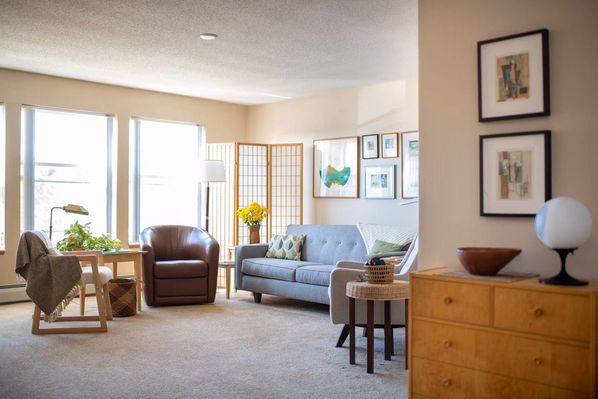 Sunlit living room with a gray sofa, brown leather armchair, wooden side tables, houseplants and framed artwork.