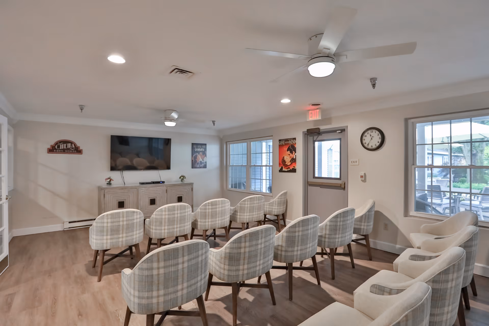 A small theater-style room with two rows of plaid upholstered chairs facing a wall-mounted flat-screen TV. The room has light-colored walls, wood flooring, ceiling fans with lights, and several windows letting in natural light. There are movie posters on the walls and a clock above an exit door, with an outdoor patio visible through the windows.