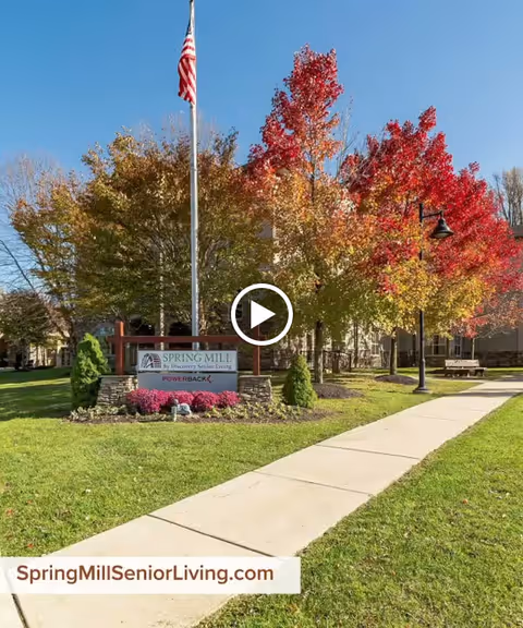 Outdoor view of Spring Mill senior living facility with a sidewalk leading to the entrance, an American flag on a flagpole, colorful autumn trees with red and yellow leaves, and a sign that reads 'Spring Mill by Discovery Senior Living POWERBACK'.