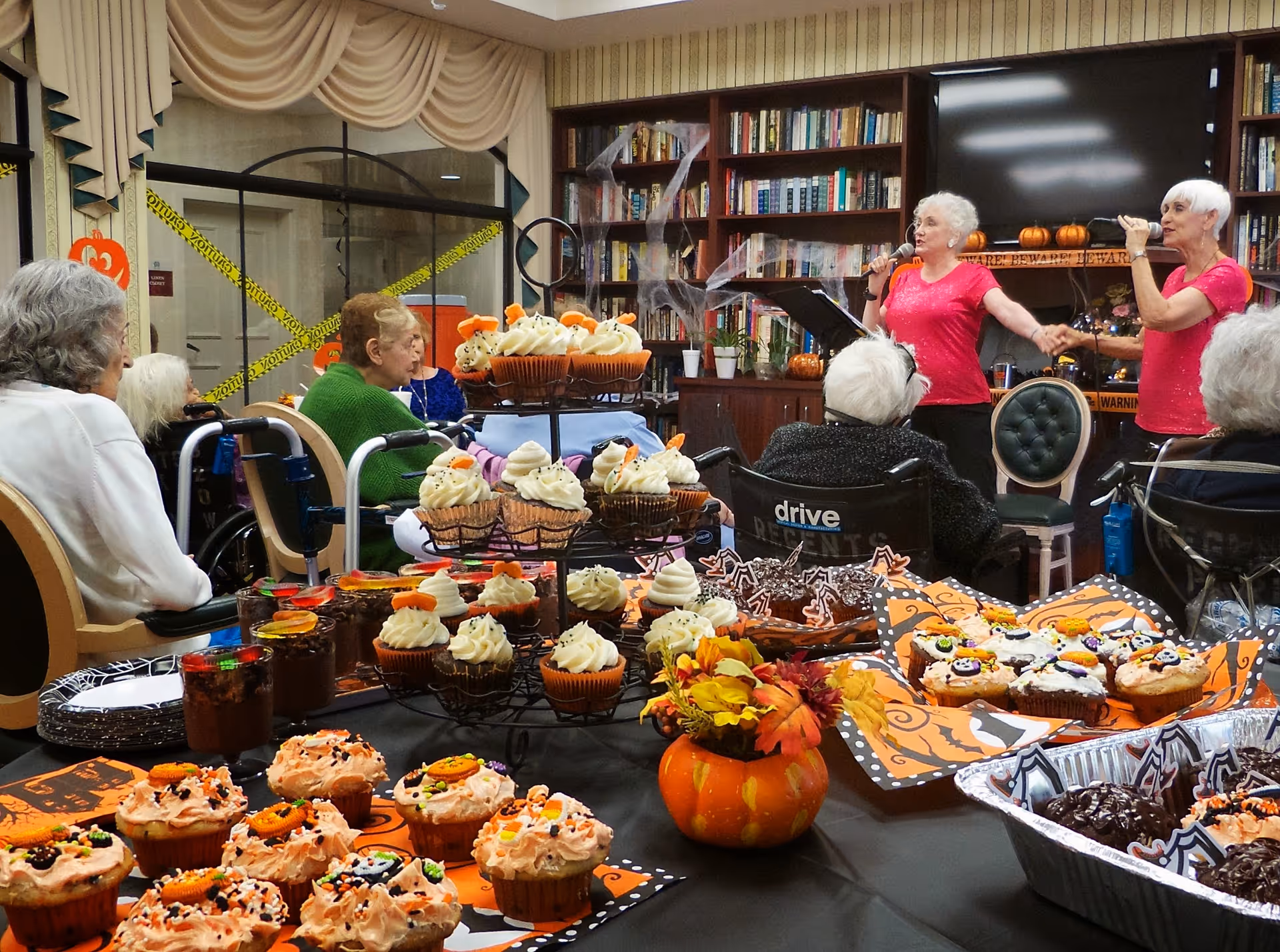 Elderly residents watch two singers while a table of Halloween-themed cupcakes and desserts fills the foreground in a library-style common room.