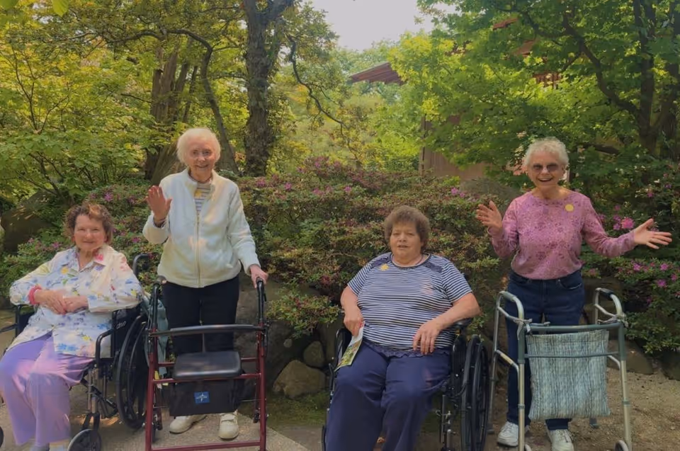 Four elderly women outdoors in a garden setting. Two women are seated in wheelchairs, one woman is standing with a walker, and another woman is standing with a rollator. They are surrounded by green trees and bushes with some flowers, and two of the women are waving and smiling.