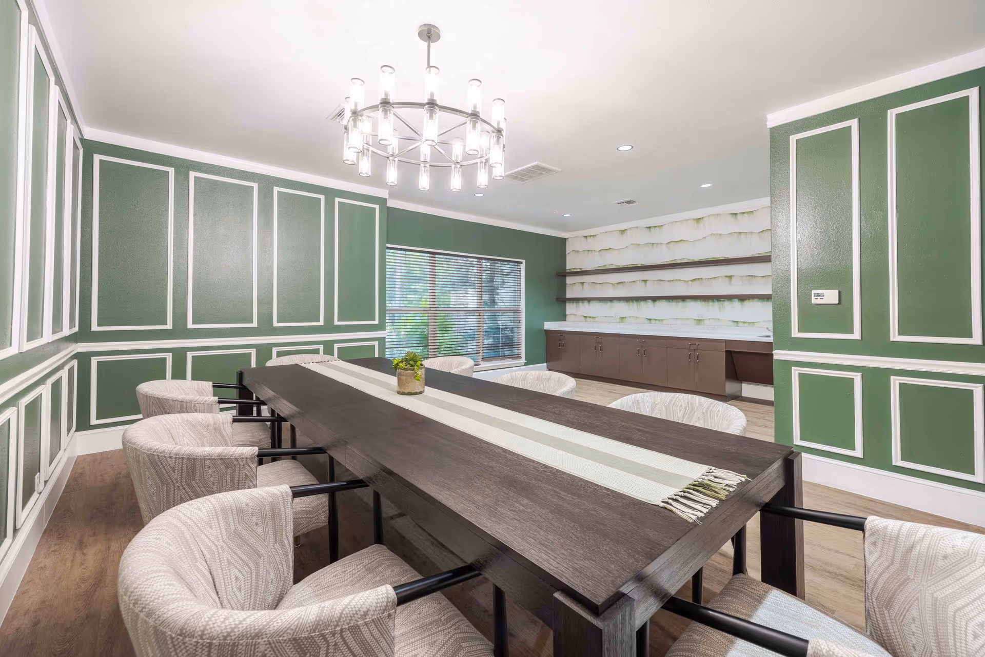 A modern dining room with a long dark wooden table surrounded by eight cushioned chairs with light patterned upholstery. The walls are painted green with white trim accents, and there is a large window with blinds letting in natural light. A contemporary chandelier hangs from the ceiling, and there are built-in shelves and cabinets along one wall.