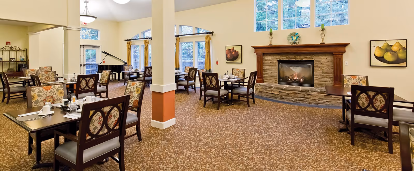 A spacious dining room with multiple tables and chairs arranged neatly. The room features a stone fireplace with a wooden mantle, decorative paintings on the walls, large windows with yellow curtains, and a grand piano in the corner. The carpet has a patterned design and the room is well-lit with ceiling lights.