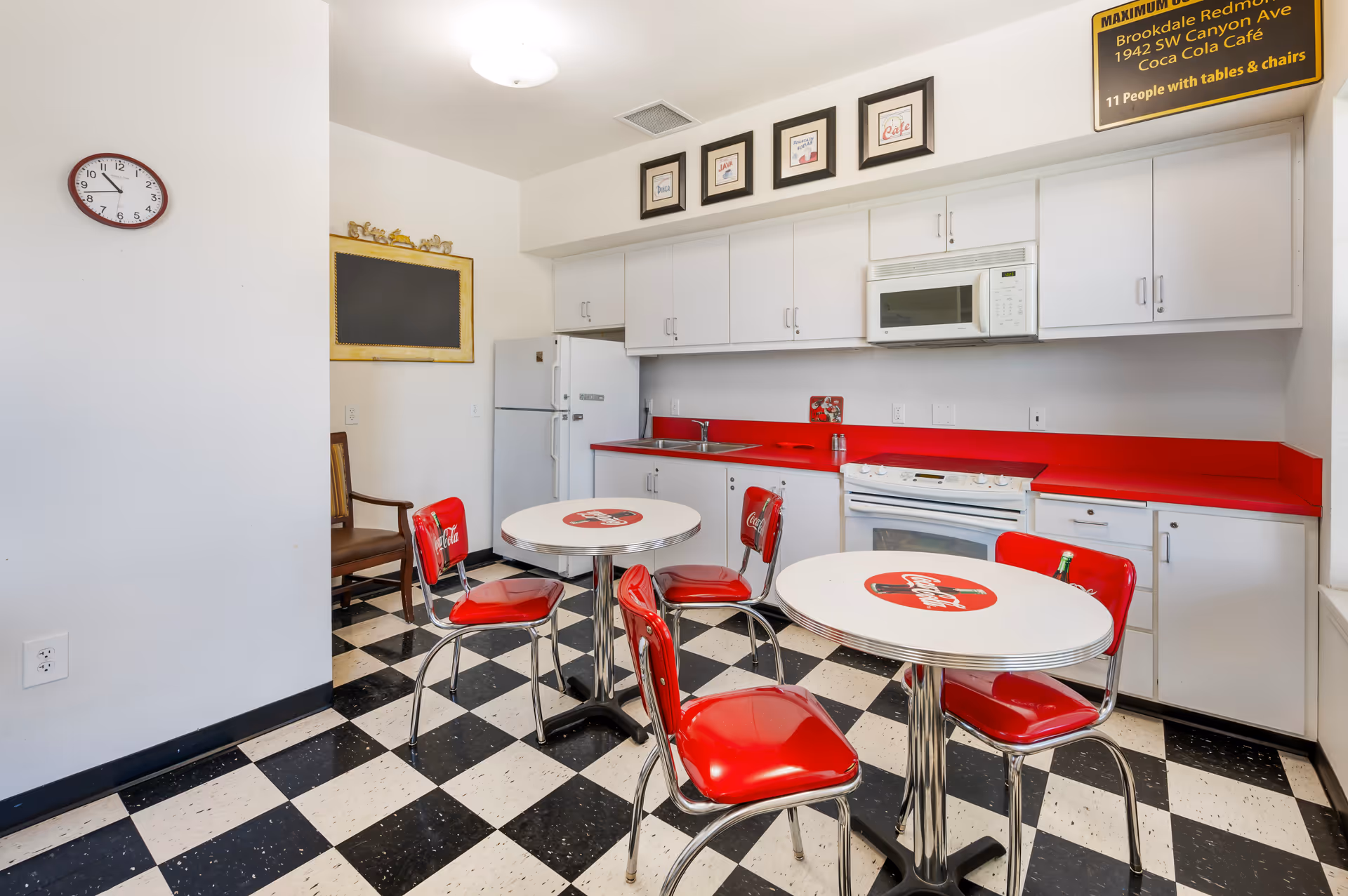 A retro-style kitchen and dining area with black and white checkered flooring, white cabinets, and red countertops. There are two round tables with Coca-Cola logos on the tabletops, each surrounded by red chairs with chrome legs. A white refrigerator, microwave, and stove are visible along the back wall. A clock is mounted on the left wall, and framed pictures hang above the cabinets. A yellow sign in the upper right corner indicates the maximum occupancy and location details for Brookdale Redmond Coca Cola Café.