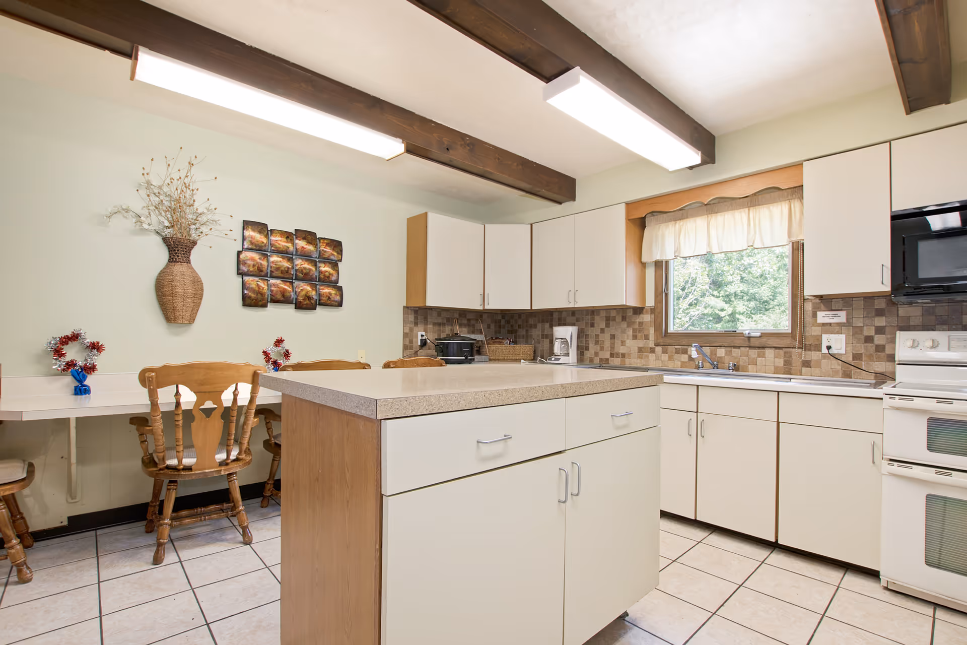 A bright kitchen with white cabinets and a beige countertop island in the center. There is a window with a cream-colored valance above the sink, and a white stove with a microwave above it. The walls have a brown tiled backsplash. To the left, there is a small dining area with wooden chairs and a wall decorated with a wicker vase holding dried branches and a grid of small square art pieces.