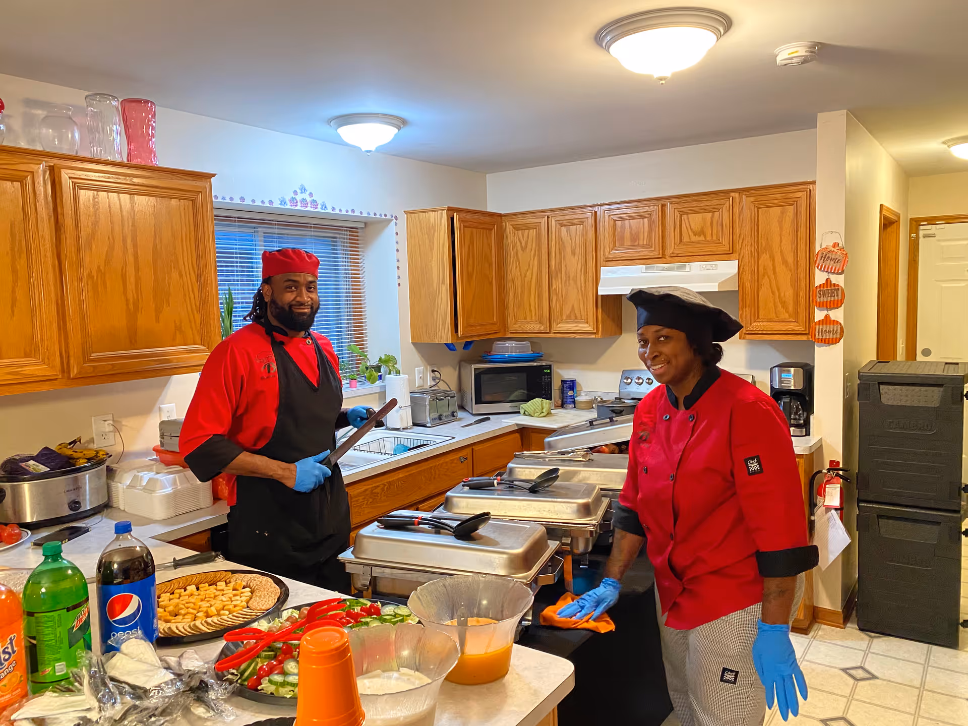 Two chefs in red uniforms and black aprons standing in a kitchen with wooden cabinets. One chef is holding a spatula and the other is wiping a surface. The kitchen counter has trays of food, plates with crackers and vegetables, and bottles of soda.