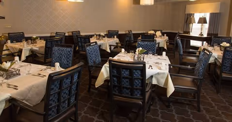 Dining room with multiple tables covered in white tablecloths, set with plates, glasses, and silverware. Dark wooden chairs with patterned blue cushions surround the tables. The room has beige walls with a subtle pattern and soft lighting from ceiling fixtures and a table lamp in the corner.