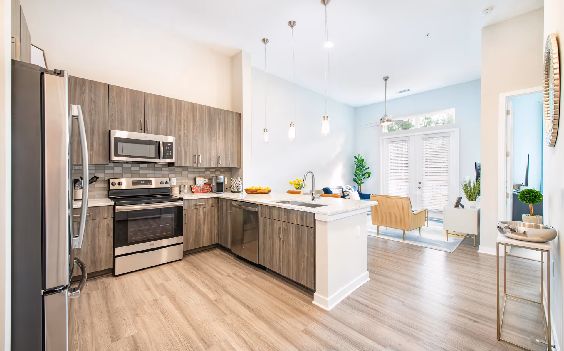 Bright modern kitchen with stainless steel appliances including refrigerator, oven, microwave, and dishwasher. The kitchen has wood-finish cabinets, a white countertop island with a sink, and three pendant lights hanging above. The kitchen opens into a living area with a blue couch, beige chair, and glass doors leading outside. Light wood flooring runs throughout the space.