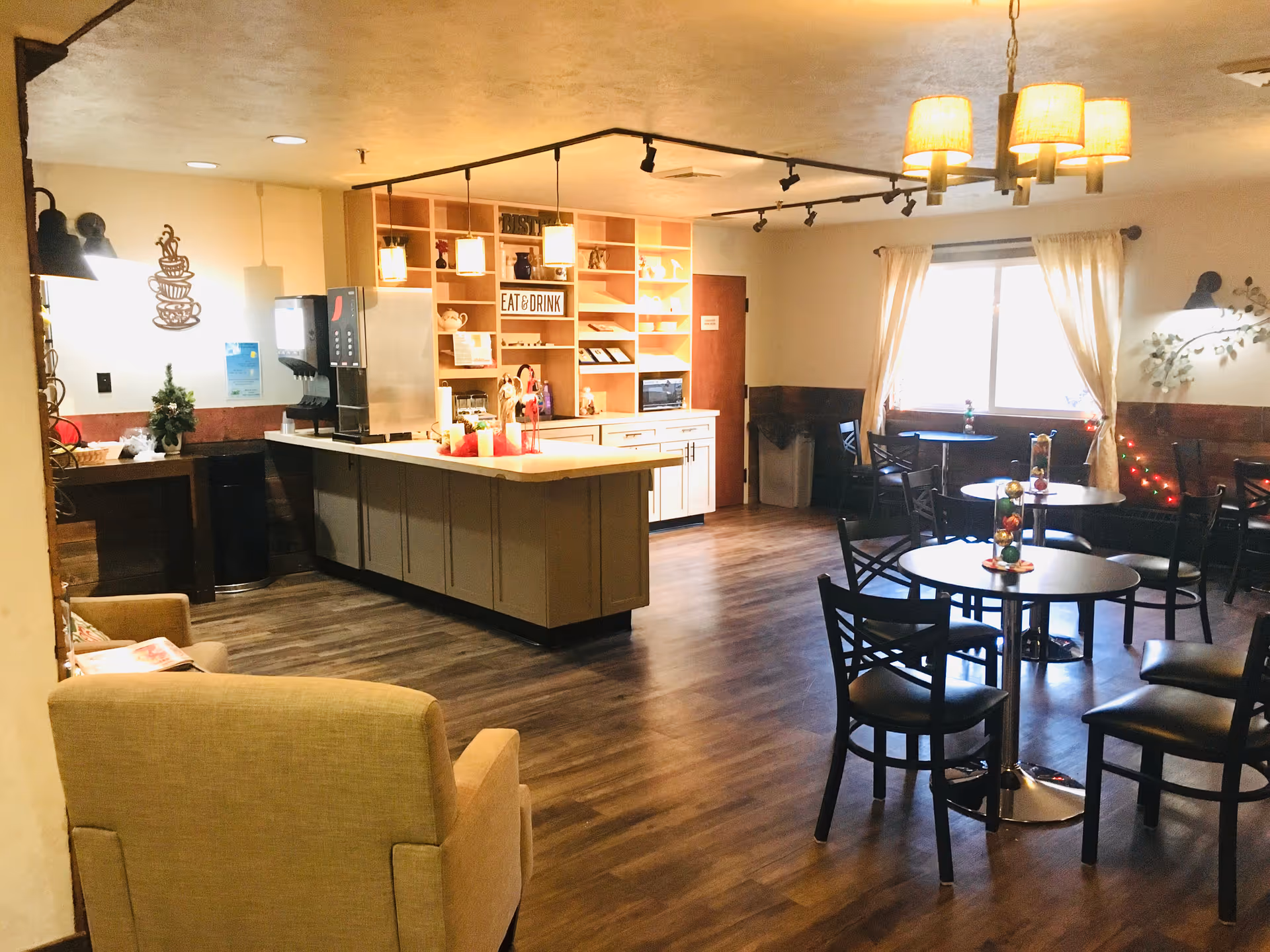 A cozy dining area in a retirement community with several round tables and chairs, a counter with a beverage dispenser, shelves with decorative items, and warm lighting fixtures. There is a window with curtains letting in natural light and a comfortable armchair in the foreground.