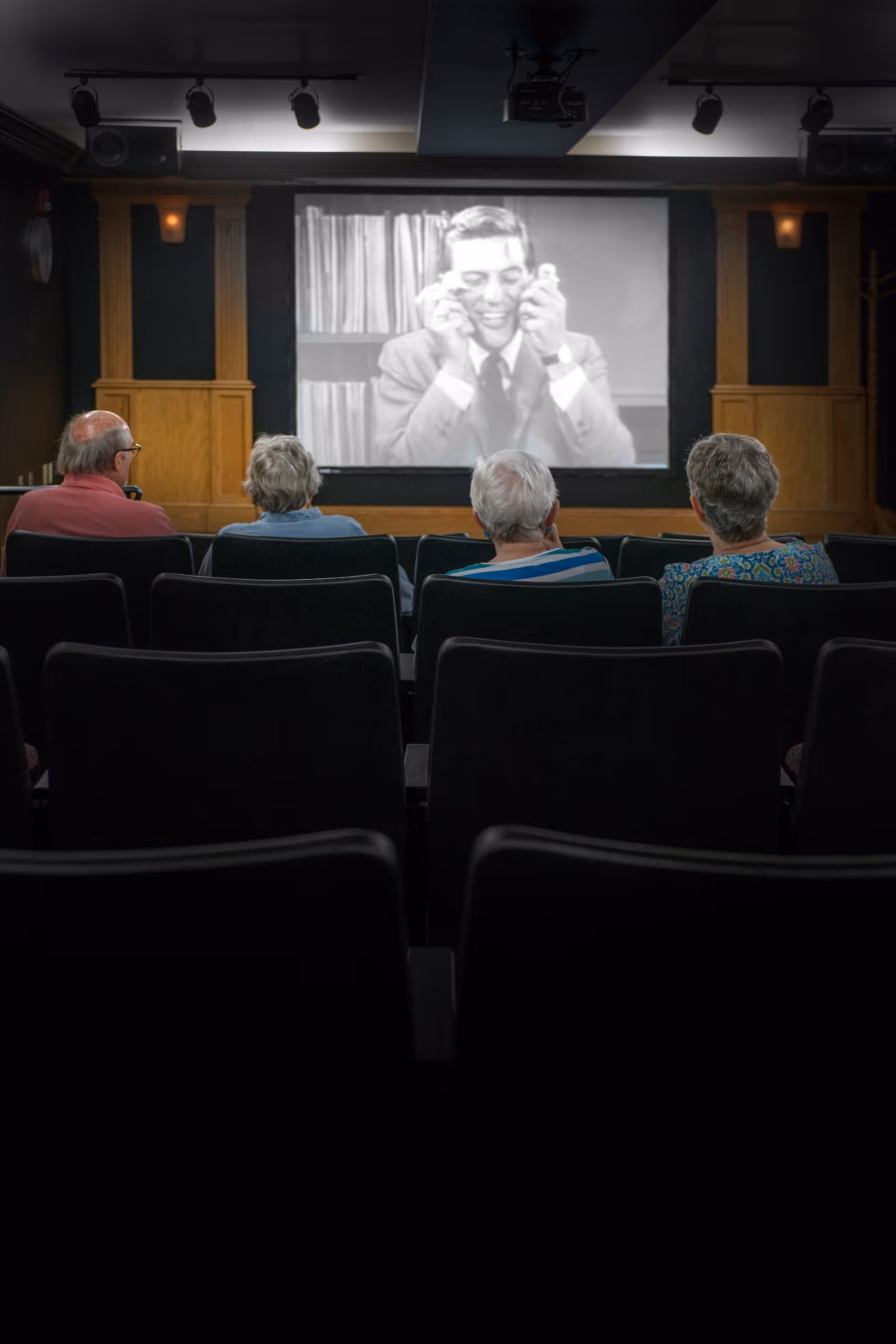 Four elderly people sitting in a small theater room watching a black and white movie projected on a screen. The room has dark walls with wooden paneling and ceiling spotlights.