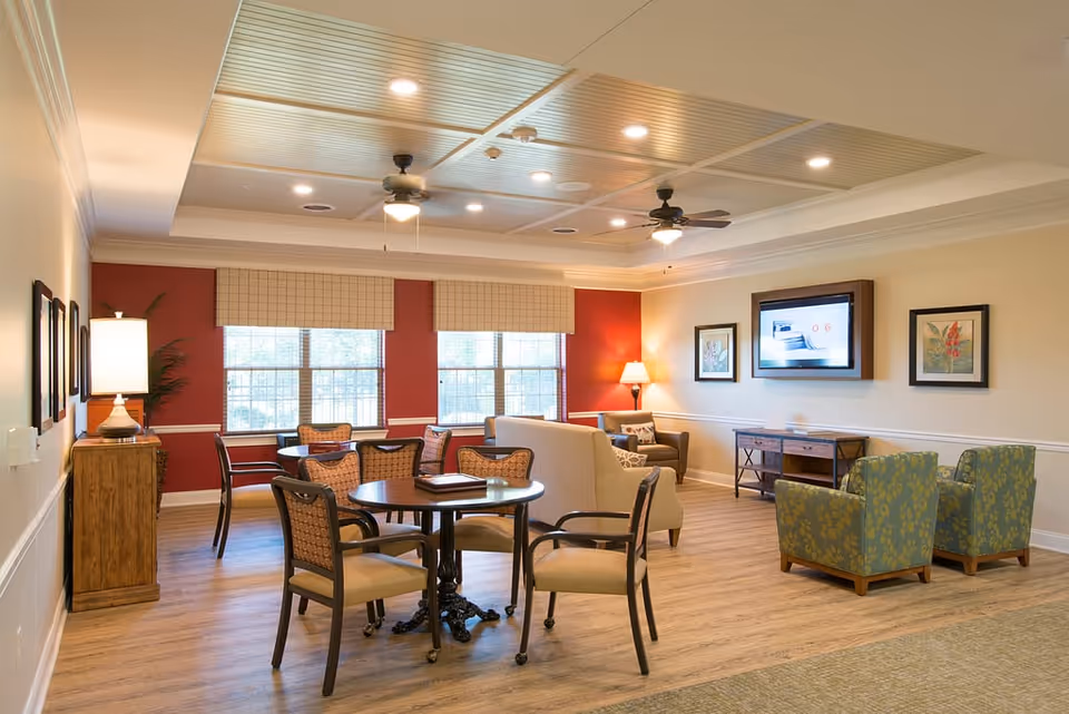 A cozy lobby area featuring a round table with chairs, a sofa, and two armchairs. The room has large windows with blinds, a ceiling fan, and decorative artwork on the walls.