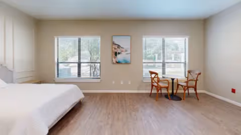 A spacious bedroom with light-colored walls and wood flooring. The room features a bed with white bedding on the left side and a small round table with two wooden chairs near two large windows with blinds. A framed picture hangs on the wall between the windows.