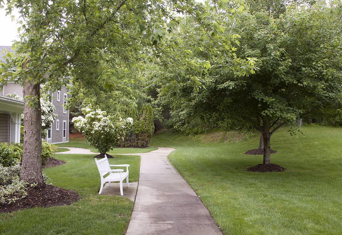 A peaceful outdoor pathway in a senior living facility with a white bench along the sidewalk, surrounded by green grass, trees, and shrubs. Part of the building is visible on the left side.