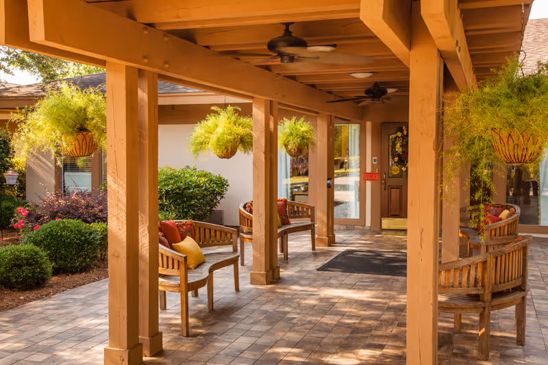 Covered outdoor entrance patio with wooden benches, hanging plants, ceiling fans and tiled floor in front of a building.