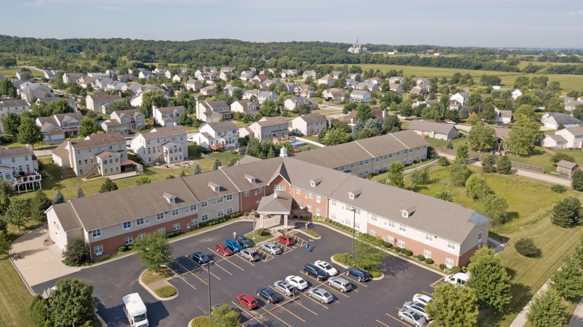 Aerial view of a two-story senior living facility with a parking lot, surrounded by suburban homes and green fields.
