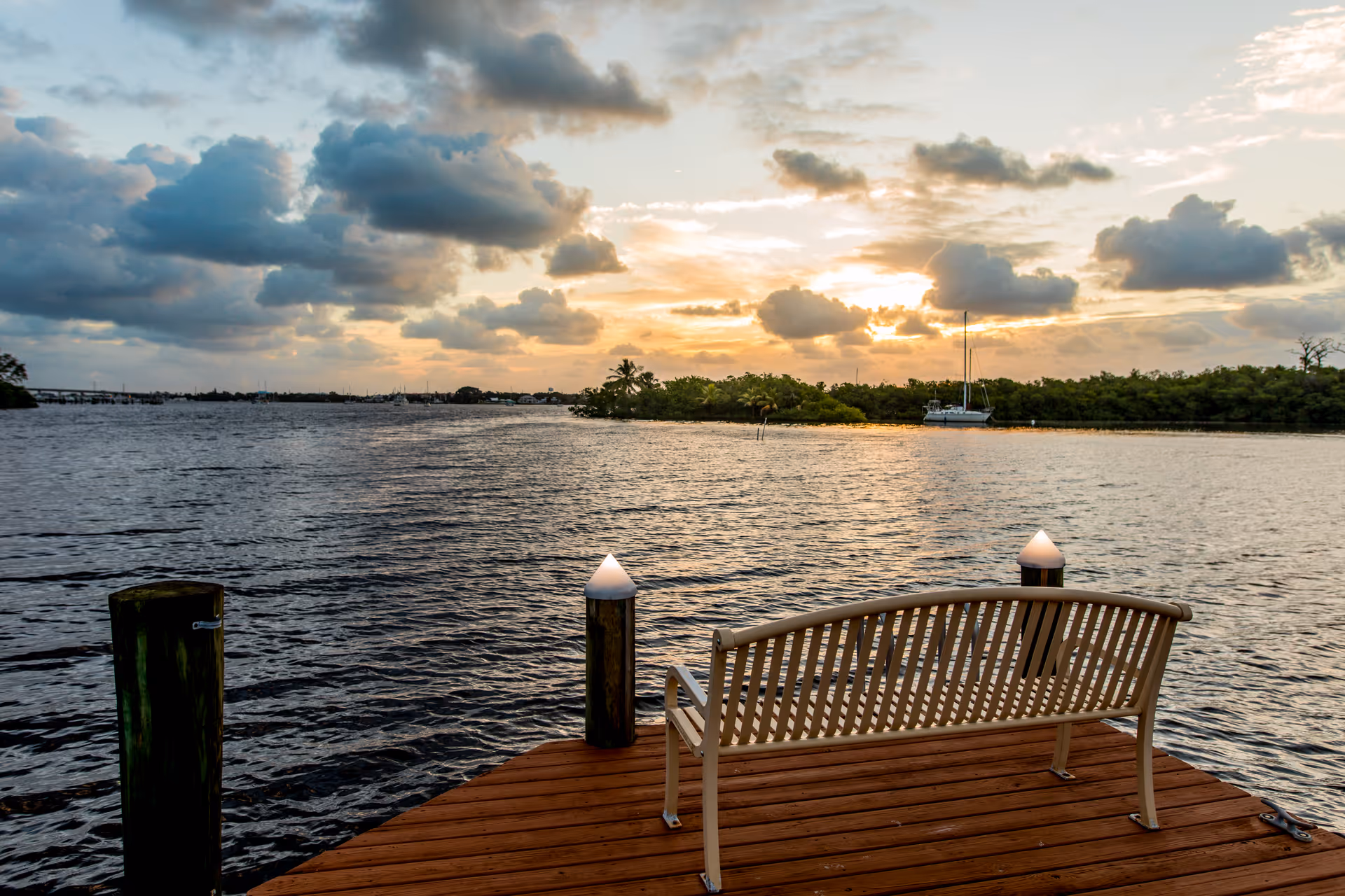 A wooden dock extending over a body of water at sunset with a bench facing the water. The sky is partly cloudy with the sun setting behind distant trees and a sailboat is visible near the shore.