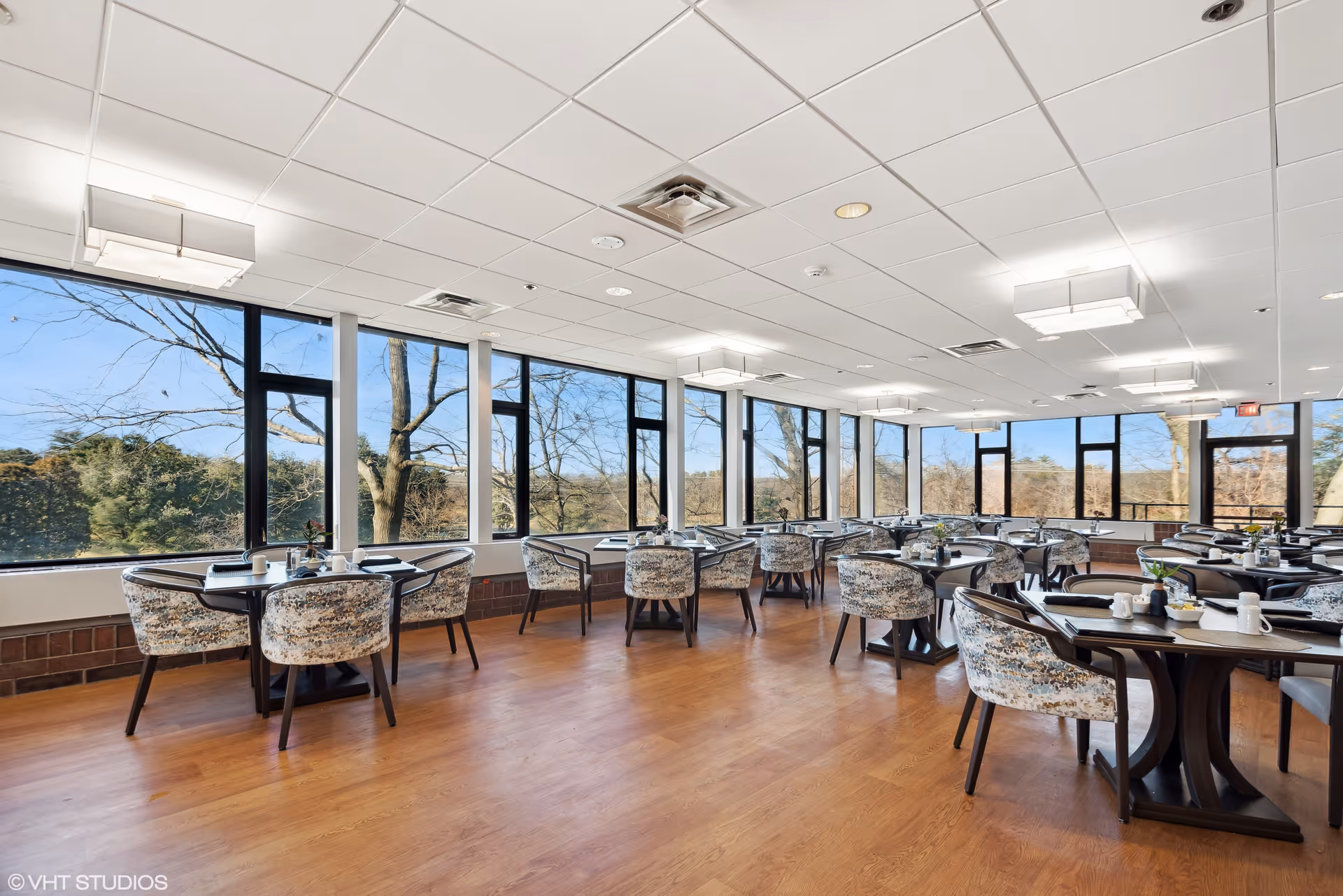 Bright dining room with large windows offering a view of trees outside. The room has multiple tables with patterned chairs arranged neatly, each table set with placemats, cups, and small decorative plants. The floor is wooden, and the ceiling has modern light fixtures.