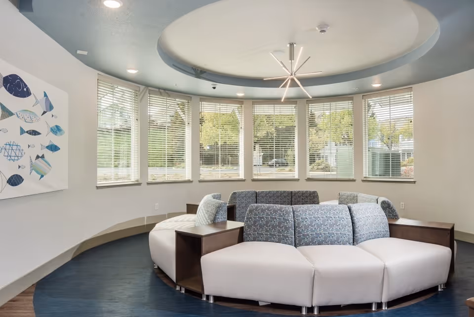 A modern circular seating area with cushioned benches arranged in a semi-circle under a circular recessed ceiling with a contemporary light fixture. Large windows with blinds surround the space, allowing natural light to enter. A wall art piece featuring various blue fish is visible on the left wall.