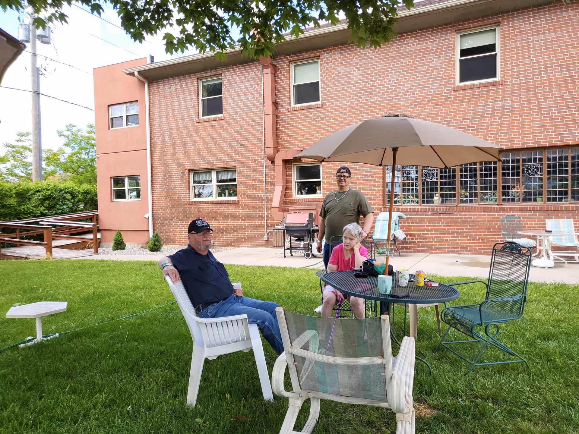 Three people sitting and standing around a patio table with an umbrella in a grassy outdoor area next to a brick building. There are various chairs around the table, and a barbecue grill is visible near the building.
