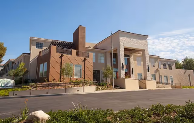 Front exterior of a two-story beige stucco senior living building with a covered entrance, driveway, and landscaped grounds under a blue sky.