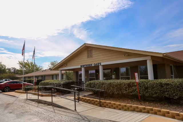 Exterior view of Northern Oaks Living & Rehabilitation Center building with a ramp leading to the entrance, bushes along the front, two flagpoles with American and Texas flags, and a clear blue sky with some clouds.