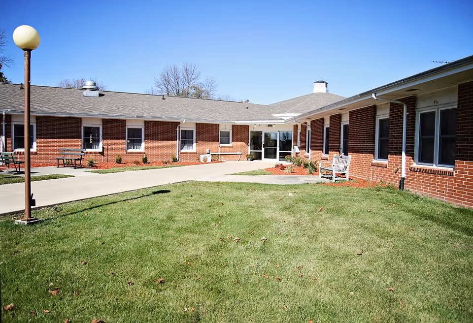 Exterior view of a single-story brick building with multiple windows and a central entrance door. The building is surrounded by a well-maintained lawn, a concrete walkway, benches, and a lamp post under a clear blue sky.