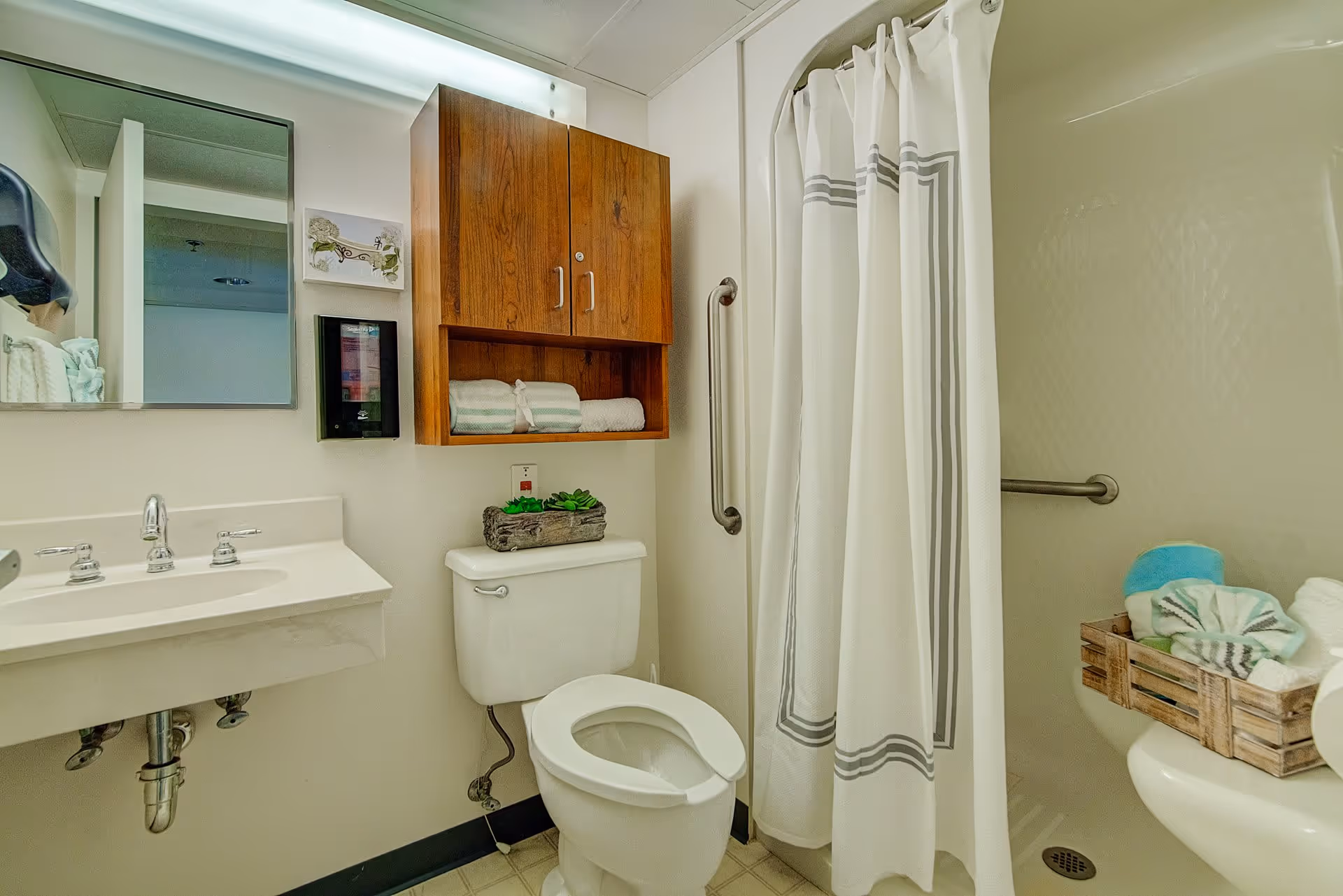 A clean bathroom featuring a white toilet with a wooden cabinet above it holding folded towels. To the left is a white sink with a mirror above it and a soap dispenser mounted on the wall. To the right is a shower area with a white curtain and a grab bar, and a wooden crate with towels on a small shelf.