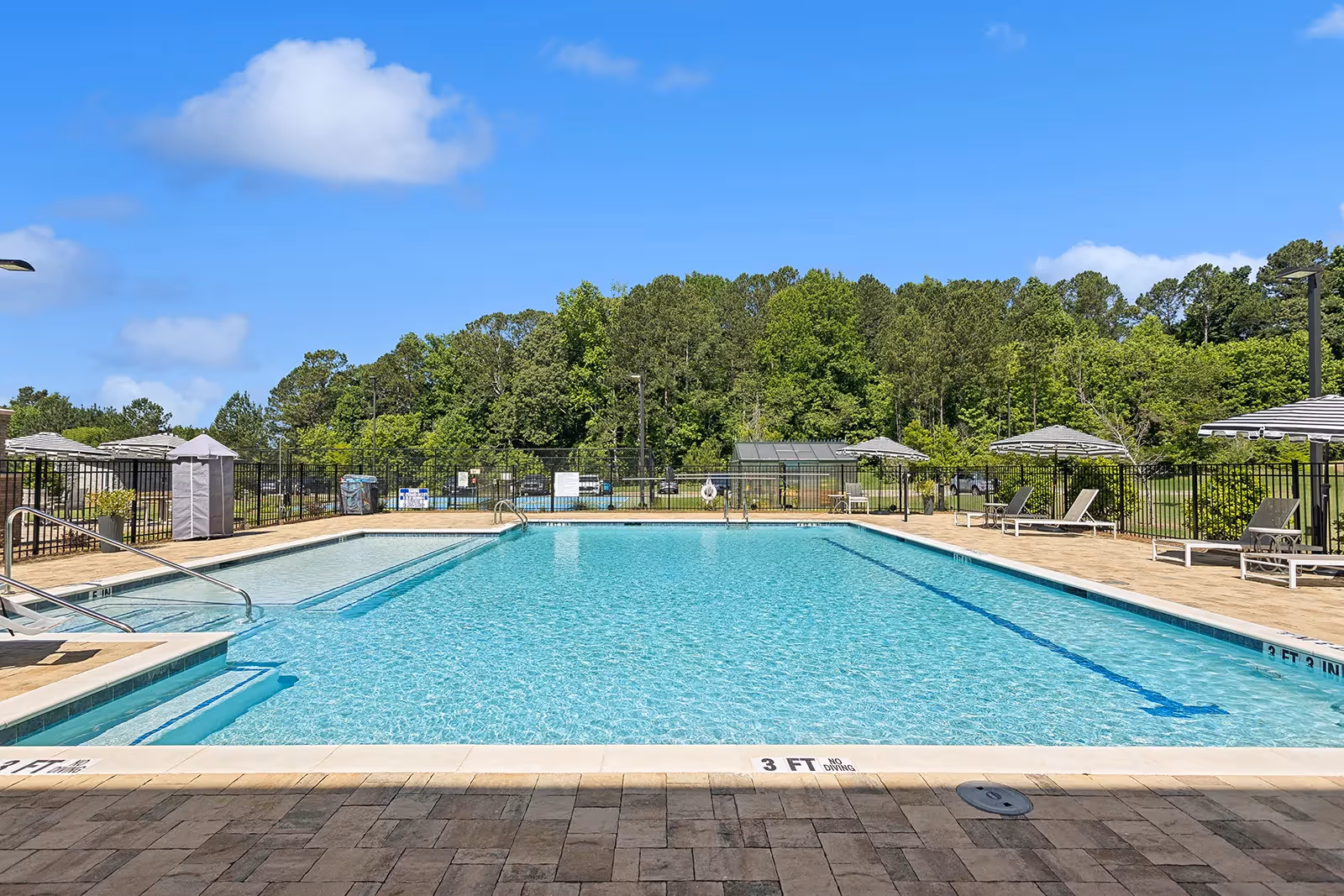Outdoor swimming pool with clear blue water surrounded by a paved deck. Several lounge chairs with umbrellas are placed around the pool area. There is a black metal fence enclosing the pool, and green trees and a blue sky with some clouds are visible in the background.