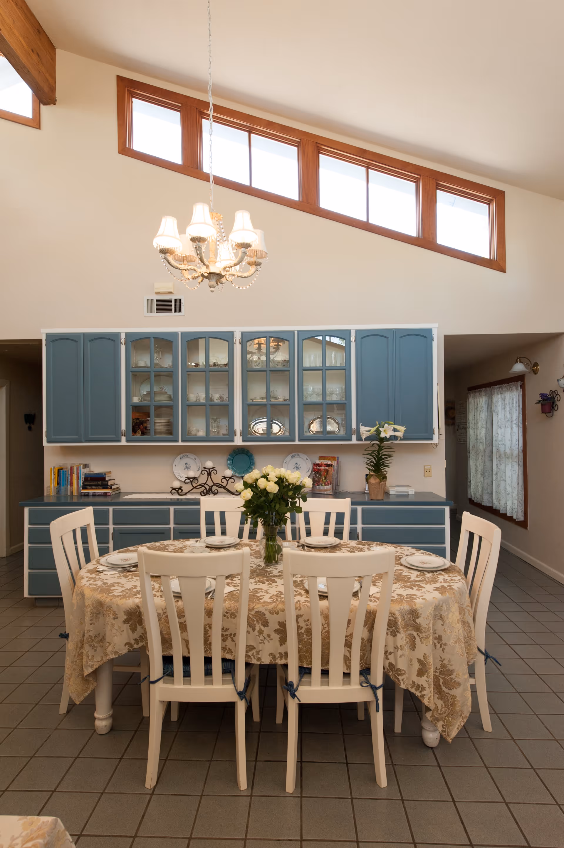 A dining area with a rectangular table covered with a floral tablecloth and set with plates and a vase of white flowers. Six white wooden chairs surround the table. Behind the table is a blue cabinet with glass doors displaying dishes and decorative items. Above the cabinet is a chandelier with five lampshades, and high windows near the ceiling allow natural light into the room.
