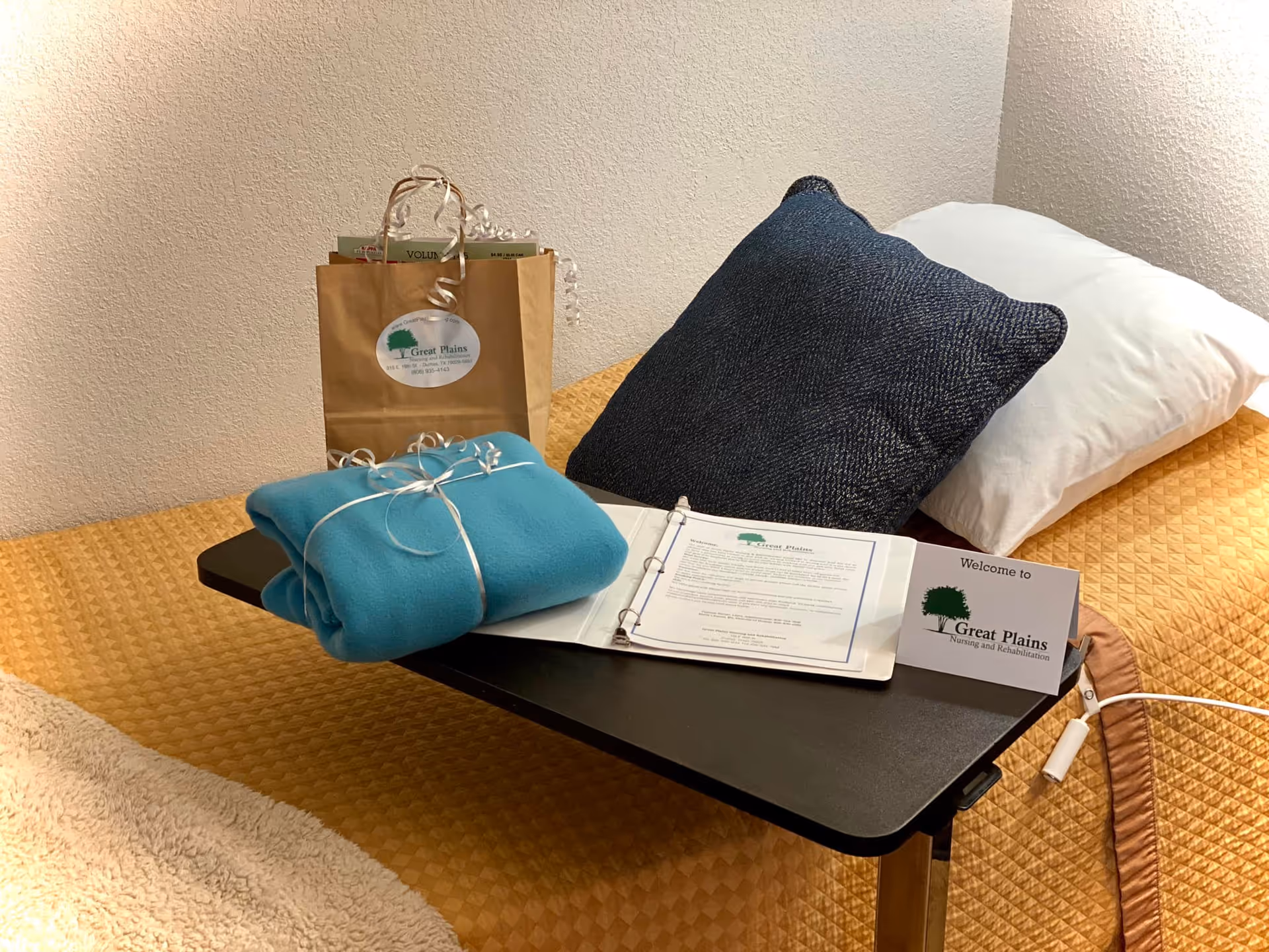 A neatly made bed with a mustard yellow quilt, a white pillow, and a dark blue decorative pillow. On the bed is a black overbed table holding a folded blue blanket tied with white ribbon, an open binder with papers, and a welcome card from Great Plains Nursing and Rehabilitation. Behind the blanket is a brown paper gift bag with the Great Plains logo and some items inside.