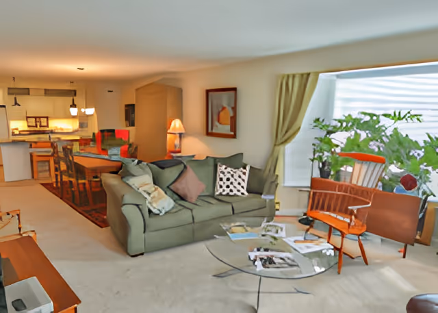 A cozy living room with a green sofa adorned with multiple pillows, a glass coffee table with magazines on it, and a wooden chair with an orange cushion. There are large windows with light green curtains and several green plants near the window. In the background, a dining area with a wooden dining table and chairs is visible, along with a kitchen area illuminated by hanging lights.