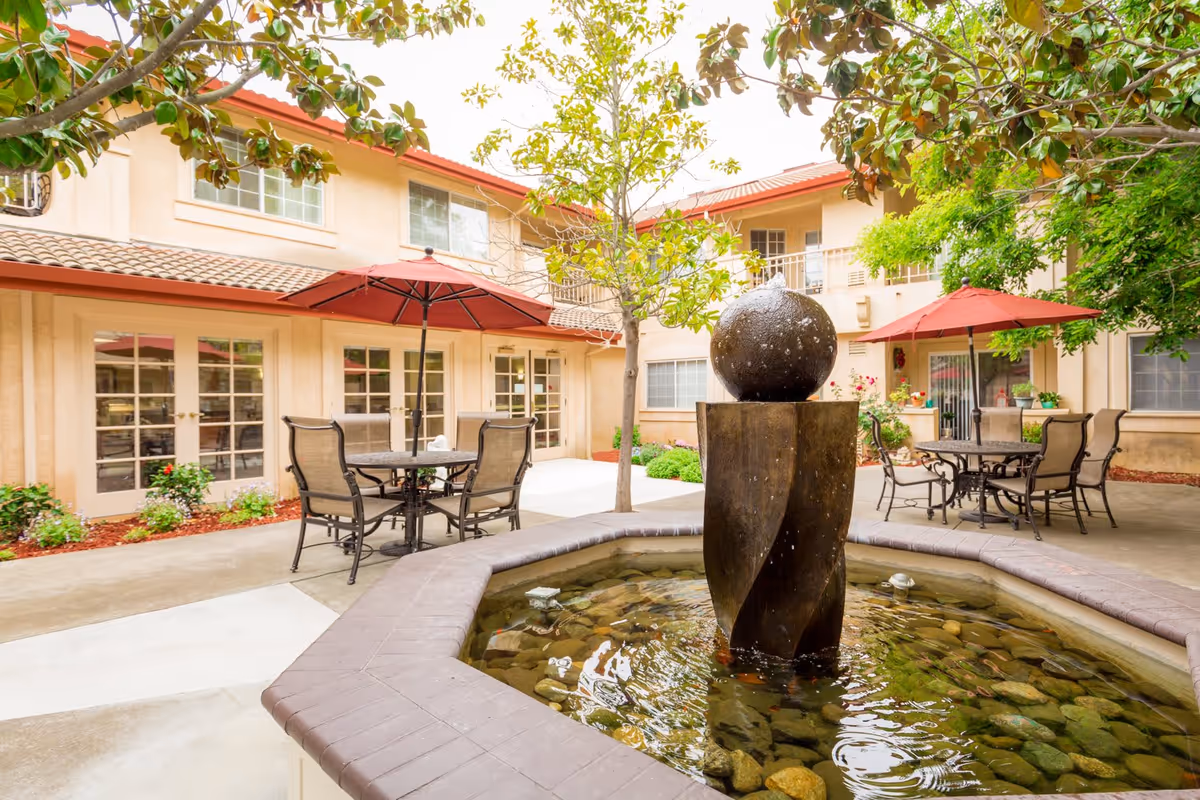 Outdoor courtyard area at Cogir of North Bay Senior Living featuring a central water fountain with a spherical top, surrounded by a stone basin filled with water and rocks. There are patio tables with red umbrellas and chairs arranged around the courtyard, with trees and plants adding greenery. The building exterior has beige walls and multiple windows and doors.