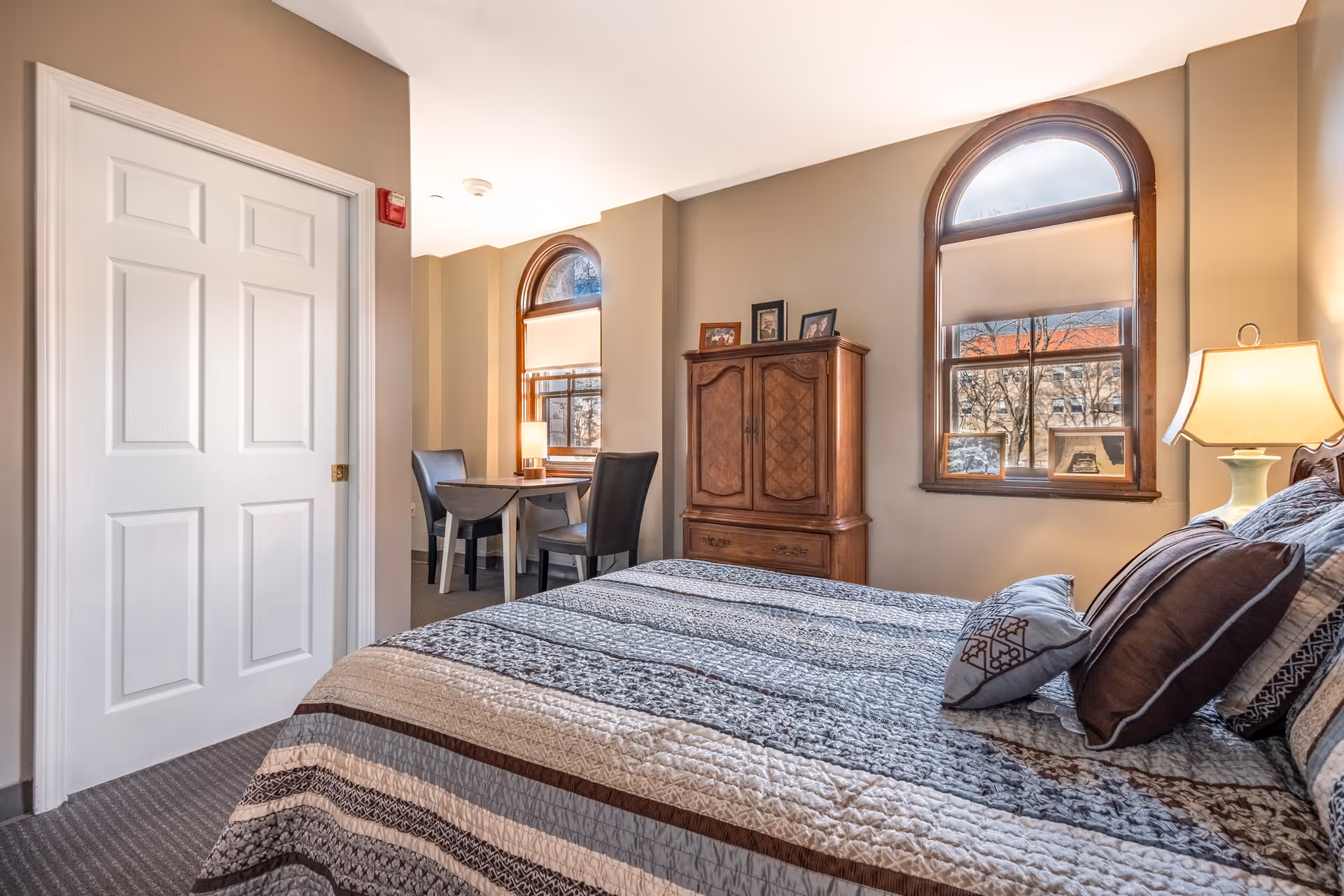 A cozy bedroom in a senior living facility featuring a neatly made bed with patterned bedding and multiple pillows. The room has two arched windows with beige blinds, a wooden cabinet with framed photos on top, a small table with two chairs near the window, and a lamp on a bedside table providing warm lighting.