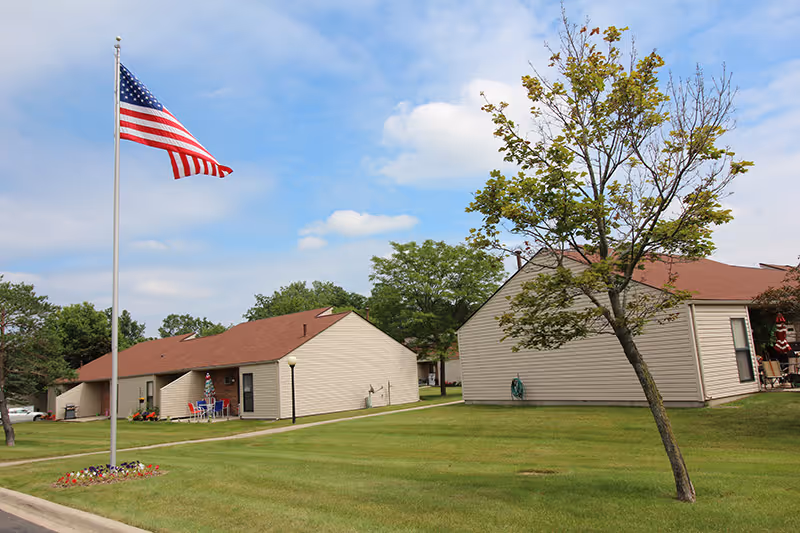 View of a senior living community with single-story beige buildings with red roofs, a green lawn, a small tree, and an American flag on a flagpole under a partly cloudy sky.