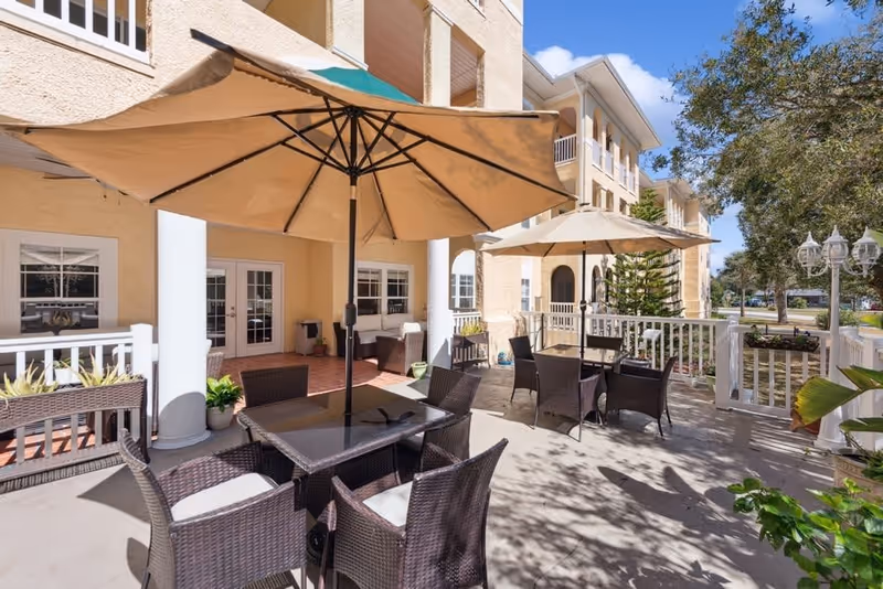 Outdoor patio area at Riviera Senior Living with multiple tables and chairs made of dark wicker, each table shaded by large beige umbrellas. The patio is adjacent to a light-colored building with white columns and railings, surrounded by greenery and trees under a clear blue sky.