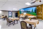 A dining room with several tables covered with beige tablecloths and chairs with patterned upholstery. The room has a mural on one wall depicting a scenic outdoor landscape with trees, mountains, and a blue sky. The floor is tiled, and there is a door and windows on the left side of the room.