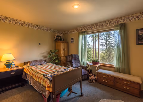 A cozy assisted-living bedroom with a single bed, nightstand, armchair, dresser, and a large window overlooking trees.