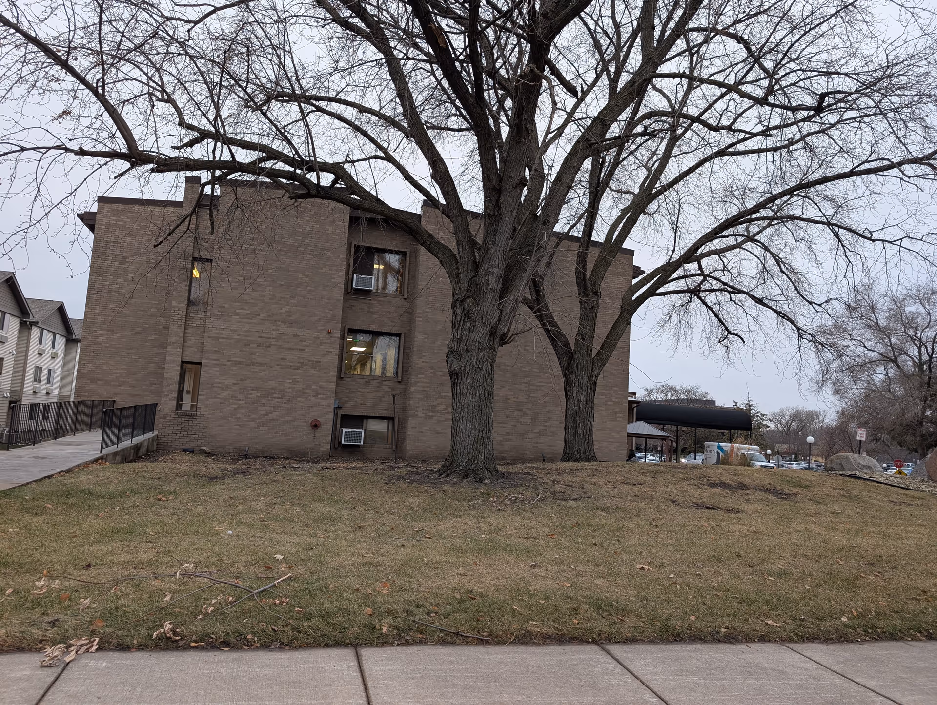 Exterior view of a three-story brick building with several windows, two large leafless trees in front, a grassy area, and a sidewalk in the foreground under an overcast sky.
