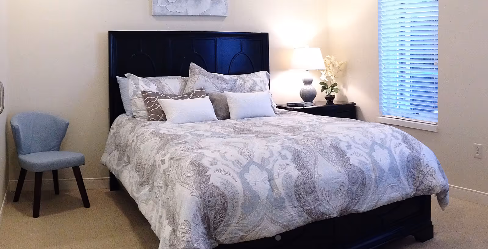 A neatly made bed with a dark wooden headboard, adorned with multiple decorative pillows and a patterned comforter in shades of white and gray. To the right of the bed is a nightstand with a lamp and a small plant. A window with blinds is visible on the right wall, and a light blue upholstered chair is positioned to the left of the bed against a light-colored wall.