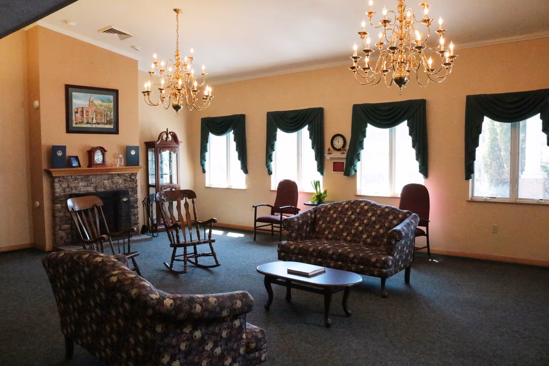 A cozy living room area with floral patterned sofas, wooden rocking chairs, and a coffee table. The room features a stone fireplace with clocks and framed pictures on the mantel, three large windows with green valances, and two ornate chandeliers hanging from the ceiling.